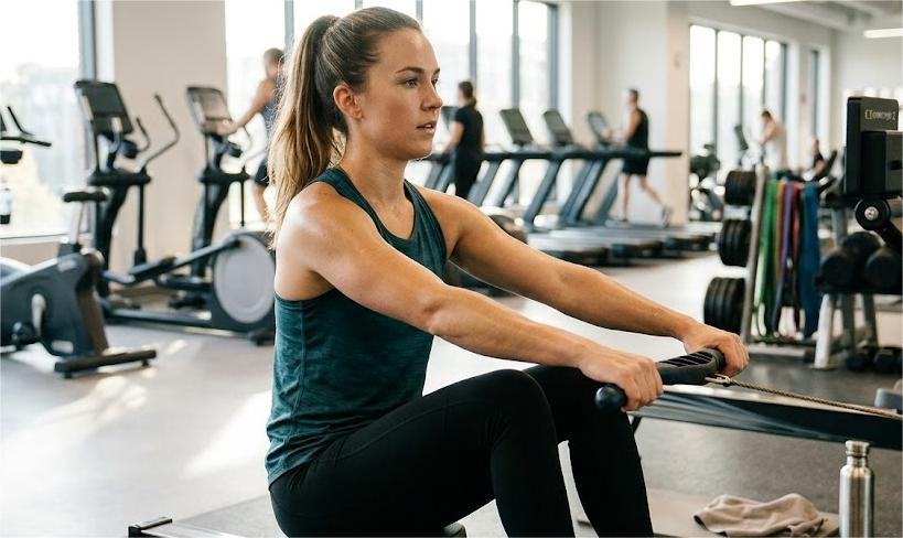 A woman exercises on a rowing machine in a bright gym, with treadmills and other gym equipment in the background.